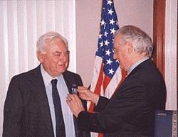 Cong. Pascrell pinning the Distinguished Flying Cross to the jacket of David Nagel in front of an American flag.