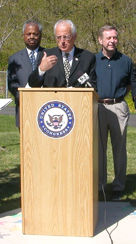 Pascrell stands at a podium in a park and is flanked by South Orange Mayor, William Calabrese to his left and Representative Donald Payne to his right. 