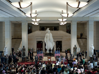 view from an upper level looking down on a large marble floor exhibition space with 19 foot white statue of a robed woman with headdress in the center, a podium and stage with people, and approximately 500 persons in the audience that are standing
