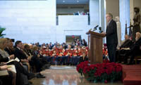 man in suit speaking on a platform at podium to a seated audience in a large marble floor exhibition space