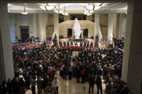 view from an upper level looking down on a large marble floor exhibition space with 19 foot white statue of a robed woman with headdress in the center, a podium and stage with people, and approximately 500 persons in the audience that are standing