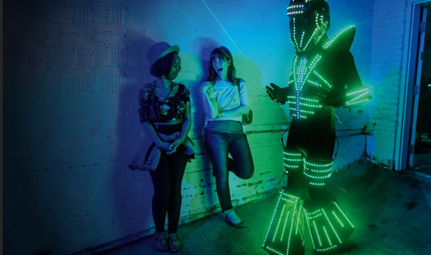 Two women in a darkened room watch a man dressed in a kind of space suit set with lights.