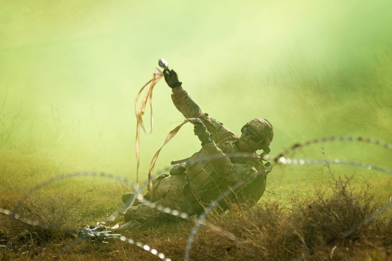 A soldier holds up objects while on the ground surrounded in smoke and concertina wire.
