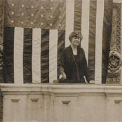 Black and white image of a Congresswoman standing in front of U.S. flag
