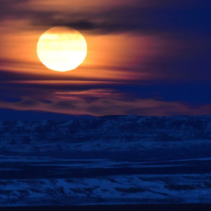 A huge full moon glows in the sky above a snowy, hilly landscape.