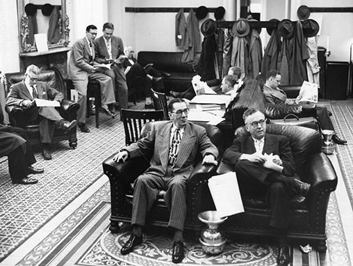 Senate Press Gallery Office filled with men and jackets & Fidora hats hanging in the background