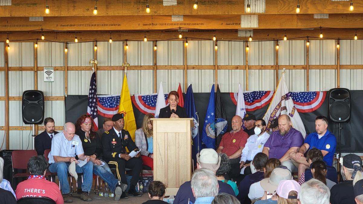 Melanie Stansbury speaks at a podium with American flag bunting as veterans bow their heads behind her.